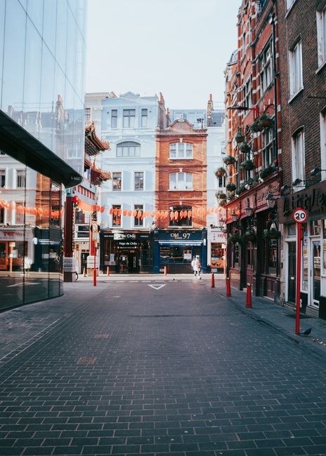 A narrow urban street scene showing a paved pedestrian area surrounded by tall, multi-storey buildings with brick and glass facades. On the right side, there are shops with large windows and signs, some with decorative hanging plants on the upper windows. The left side features reflective glass storefronts, reflecting the opposite buildings. In the background, a mix of historical and modern architecture is visible, with a clear, bright sky overhead. String lights with orange bulbs stretch across the street, indicating a festive or lively atmosphere. Red bollards line the sidewalk, and a 20 mph speed limit sign is attached to a pole on the right. The scene is well-lit, capturing the clean and orderly environment typical of a city centre, and subtly relates to services such as private rubbish removal or on-site clearance handling waste from commercial properties in urban areas, reflecting the context of alternative waste management options.