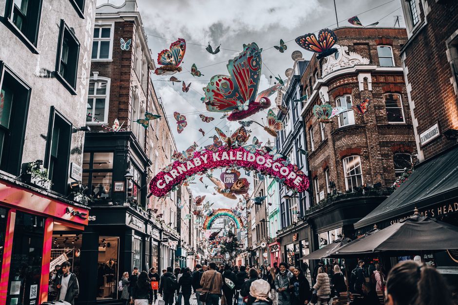 Wide-angle view of Carnaby Street in Soho during evening hours, featuring an illuminated, multicoloured sign arching across the street spelling 'CARNABY' in individual letters with a vintage-style white sign underneath. The scene captures brick facades of buildings on both sides, with warm lighting emanating from large window displays on the left, revealing indoor decor and potted plants on window sills. On the right, a black security camera is mounted near the top corner of a building, adjacent to a street sign reading 'Kingly Street.' String lights are suspended overhead, creating a festive atmosphere against a deep blue evening sky. The buildings exhibit a mix of brickwork and painted finishes, with one featuring a dark grey edge trim. The overall scene conveys an urban environment prepared for visitor activity, with subtle hints of ongoing street life and the area's distinctive commercial character, reflecting the typical setting where private waste collection or rubbish removal services might operate in a busy city district like Soho.