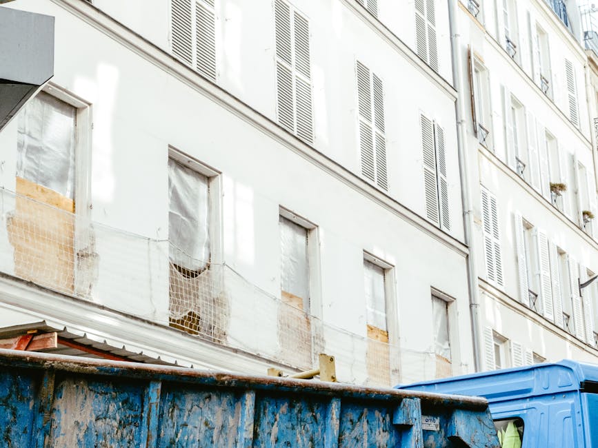 The image depicts the lower section of a multi-storey residential building with white painted exterior walls and closed window shutters, some with wooden panels. Several windows are visible, with some still boarded up or covered with protective plastic or netting, indicating ongoing renovation or maintenance work. In the foreground, a large, weathered blue skip sits on the pavement, showing signs of rust and chipped paint, used for the temporary collection of debris and waste. A portion of a green-tinted construction or waste truck is partially visible behind the skip, emphasizing the context of waste clearance or rubbish removal. The scene suggests a typical urban setting where private waste management, such as skip hire services from Rubbish Removal Soho, might be engaged as an alternative to council collection for building refurbishment or renovation projects, aligning with the topic of independent collection and on-site clearance solutions in the Soho area. The environment is lit by natural daylight, creating a clear and neutral tone consistent with professional waste management operations.