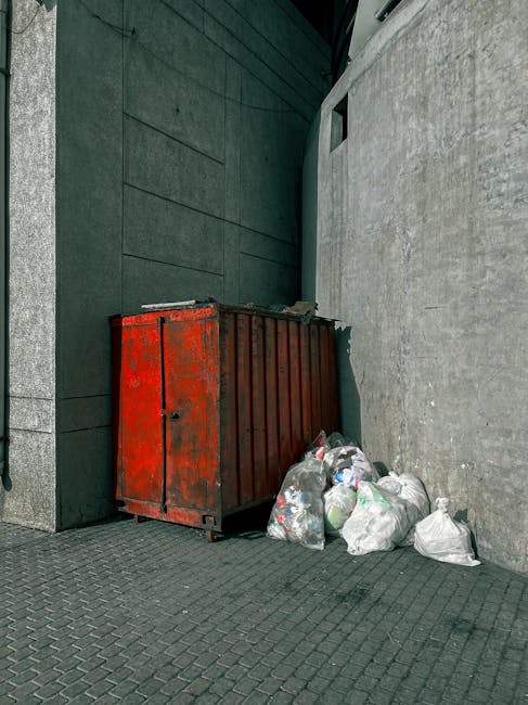 A large, rectangular metal skip painted in faded red with visible rust and chipped paint is positioned against a tall, dark green wall and a light gray concrete building exterior. Several clear plastic bags filled with waste material are placed on the cobblestone ground next to the skip, with some bags partially obscuring the base of the container. The skip appears to be in an urban alleyway or service area, with a textured paving surface beneath and around it. The surrounding environment includes the dark green wall on the left, the concrete wall on the right, and a small, dark rectangular opening near the top of the concrete structure. The scene is evenly lit with natural daylight, emphasizing the utilitarian setting for private waste handling or alternative rubbish removal services conducted by Rubbish Removal Soho in the context of non-standard waste collection options in the W1 postcode area.