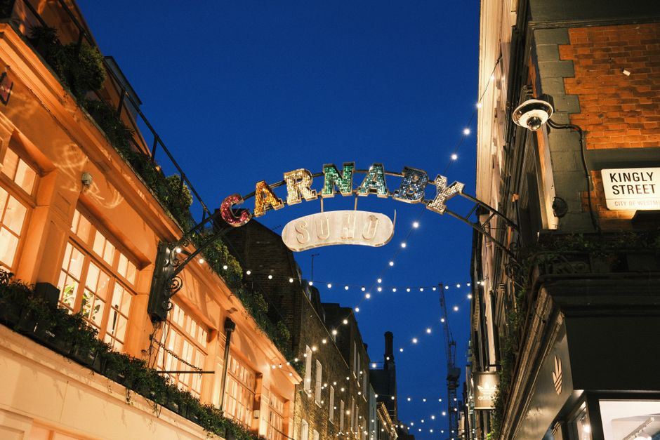 A night-time street scene in Soho featuring a decorative archway sign reading 'CARNABY' illuminated with colorful, vintage-style metal letters, some of which are lit with small bulbs, and a smaller sign below reading 'SOLD' in metal. Surrounding buildings are constructed from brick and plaster, with large windows framed by wooden trim. The left side shows a building with illuminated windows and balcony planters filled with greenery, while the right side has a black sign for a business and exterior lighting fixtures attached to the brick facade. String lights are suspended above the street, creating a warm, festive atmosphere against the dark blue evening sky. The scene is captured from street level, emphasizing the architectural details and decorative signage, subtly linking to the theme of vibrant city nightlife and private areas that may require occasional rubbish removal or maintenance services, such as those provided by Rubbish Removal Soho.