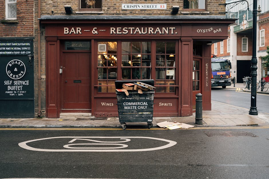 In front of a brick-walled restaurant on Dean Street Soho, a black commercial waste bin labeled 'COMMERCIAL WASTE ONLY' is positioned on the pavement. The bin is partially open, revealing scattered cardboard boxes and waste paper inside, with some discarded papers lying on the ground nearby. The restaurant's large front windows have wooden framing and display warm interior lighting, with a maroon exterior finish accentuating the facade. To the right of the bin, a short metal post and a bollard are visible, aiding pedestrian separation from the road. Behind the bin, a parked truck is partially seen on the street, and the adjacent buildings include a bicycle rack and a street sign above indicating 'Crispin Street.' The scene captures a typical urban environment where private waste collection may be taking place, aligning with commercial rubbish removal services such as those offered by Rubbish Removal Soho, situated amidst local businesses and city infrastructure on a relatively quiet street.